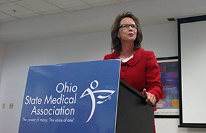 Image of a  woman wearing a red suit jacket speaking from a podium.