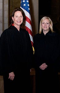 Image of two female judges wearing black judicial robes standing side-by-side in front of the American flag.