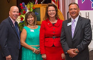 Image of two men wearing dark suits standing on either side of two women, one wearing a green dress and one wearing a red dress.