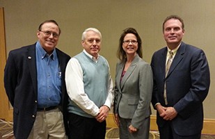 Image of a woman wearing a grey suit standing with three men, two wearing dark suits and one wearing a white dress shirt and sweater vest.