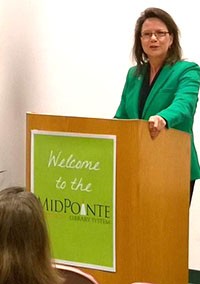 Image of a woman wearing a green suit jacket and black blouse speaking from a podium.