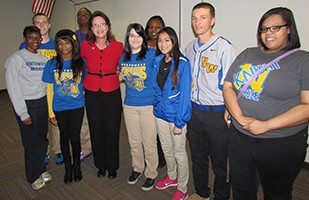 Image of a woman wearing dark dress pants and a red suit jacket standing with a group of high school students.