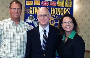 Image of two men, one wearing a plaid dress shirt and one wearing a dark suit standing next to a woman wearing a black suit and green blouse.