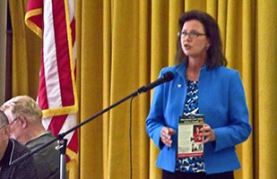 Image of a  woman wearing a wearing a blue suit standing in front of a gold curtain speaking into a microphone.