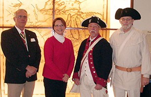 Image of a man wearing a blue suit jacket, a woman wearing a red sweater and black dress pants, and two men dressed like Revolutionary War soldiers.