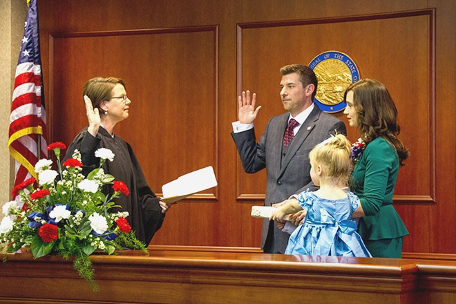 Image of a female judge wearing a black judicial robe standing with her right hand raised in front of a man wearing a grey suit, a woman in a green dress, and a female toddler wearing a blue dress.