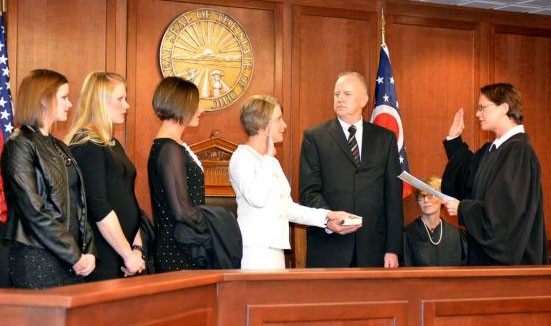 Image of a woman wearing a white dress with her right arm raised standing in front of a female judge wearing a black judicial robe. Three women, all wearing black dresses, are standing to one side, and a man wearing a black suit stands on the other side.