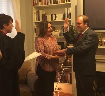 Image of a female judge wearing a black judicial robe standing in front of a man wearing a grey suit. Both have their right hands raised. a woman wearing a multi-colored blouse and dark dress pants holds a book between them.