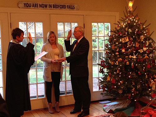 Image of a female judge wearing a black judicial robe standing in front of a man wearing a black suit. Both have their right hands raised. A woman wearing black pants, a white top and a long sweater holds a book between them.