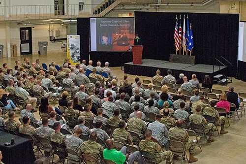 Image of a woman wearing a black suit and green blouse speaking from a podium to a room full of men and women dressed in fatigues.