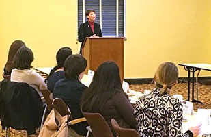 Image of a woman wearing a black suit and red blouse speaking from a podium to a room full of men and women.