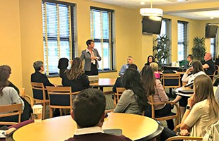 Image of a woman wearing a grey suit jacket and black pants speaking to a room full of men and women.