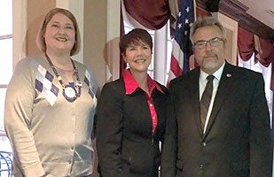 Image of a woman wearing a black suit and red blouse standing between a woman wearing a beige, argyle sweater and a man wearing a black suit.