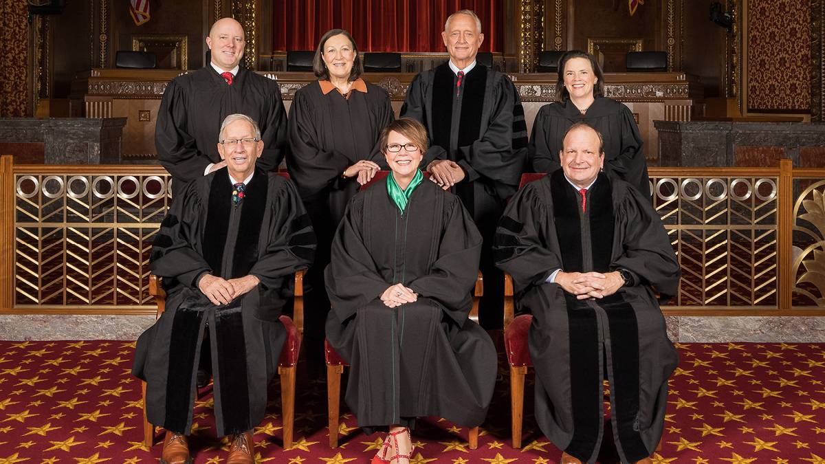 Seven men and women, four standing behind three who are seated, all wearing black judicial robes, in the courtroom of the Thomas J. Moyer Ohio Judicial Center.