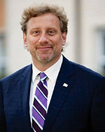 Image of a man with sandy blond hair and a close-shaved mustache wearing a navy suit and purple and navy striped tie.