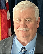 Image of a man with grey hair and a mustache wearing a grey suit and blue dress shirt.