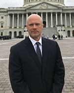 Image of a bald man wearing a dark suit and tie standing in front of a large, marble building with columns.