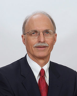 Image of a man with a grey mustache wearing a navy suit and red tie.